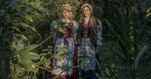 Zdjęcie przedstawia dwie młode kobiety - autorki projektu fotograficznego "Czekając na Śnieg", pozujące w kostiumach inspirowanych tradycyjnymi strojami polskimi na tle roślinności tropikalnej.