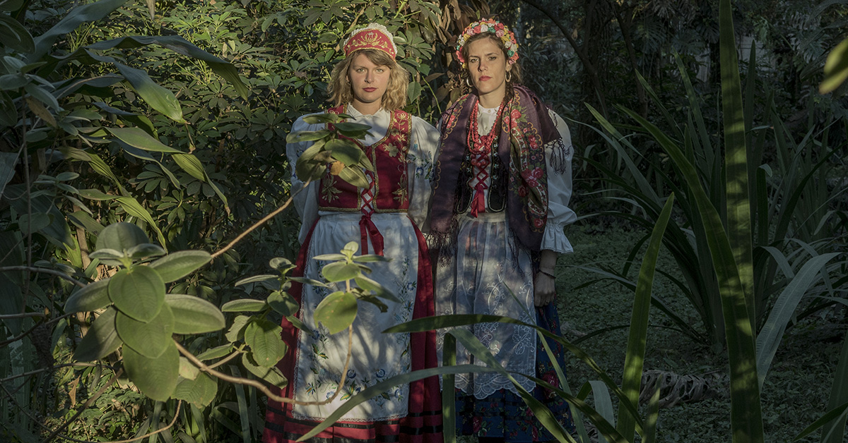 Zdjęcie przedstawia dwie młode kobiety - autorki projektu fotograficznego "Czekając na Śnieg", pozujące w kostiumach inspirowanych tradycyjnymi strojami polskimi na tle roślinności tropikalnej.