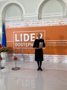 Fotografia, wnętrze pałacu, w centrum kobieta ubrana w czarną sukienkę trzyma w rękach statuetkę. W tle widoczny napis Lider Dostępności