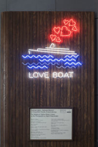 Zdjęcie. Przestrzeń wystawy Love Boat. Ściana w kolorze ciemnego drewna. Na górze ściany wisi niebiesko-biało-czerwony neon przedstawiający płynący po falach statek. Z komina statku wydostaje się dym w kształcie czerwony serc. Pod niebieskimi falami świecący napis Love Boat. Pod neonem tabliczka z tekstem kuratorskim wystawy.
