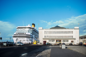Budynek Muzeum Emigracji widziany od frontu. Jasny budynek w stylu gdyńskiego modernizmu, z ciemną kopułą. Obok budynku duży statek wycieczkowy zacumowany przy Nabrzeżu Francuskim. Nieco wyższy niż budynek Muzeum. Statek jest biały, ma kilka pięter. Na górze widać żółty komin.