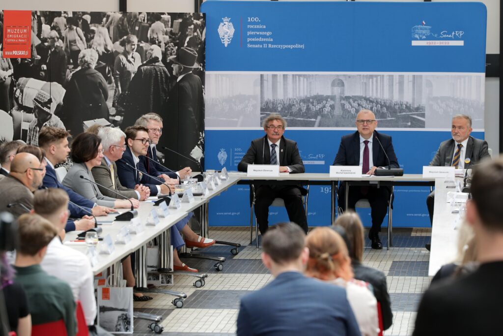 zdjęcie przedstawia członków komisji na czele z przewodniczącymi oraz uczestników spotkania w sali obrad, to jest w lobby Muzeum Emigracji na tle okazjonalnej dekoracji ściennej z logotypami Komisji Spraw Emigracji i Łączności z Polakami za Granica.