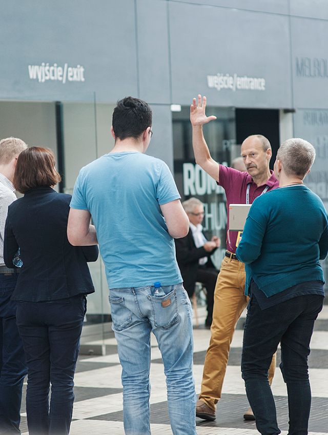 Kolorowe zdjęcie. Wolontariusz z grupą zwiedzających w holu muzeum. Wolontariusz trzyma rękę w powietrzu wskazując kierunek zwiedzania.