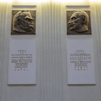 Zdjęcie. Widok na dwie pamiątkowe tablice znajdujące się nad schodami w Holu pasażerskim. To dwie płaskorzeźby wykonane z brązu przedstawiające zwrócone do siebie profile twarzy Józefa Piłsudskiego oraz Ignacego Mościckiego. Pod płaskorzeźbami złote napisy.