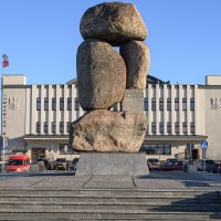 Zdjęcie. Widok na skwer im. Witolda Gombrowicza. W centralnej części zdjęcia pomnik na niskim cokole na placu wyłożonym kostką. Pomnik składa się czterech głazów ułożonych w pionową kompozycję. Na dalszym planie po prawej stronie frontowa fasada Dworca Morskiego, obecnie siedziba Muzeum Emigracji w Gdyni. Po lewej stronie zdjęcia magazyn oraz żuraw portowy.