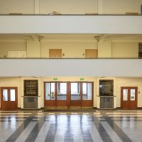Zdjęcie. Wyjście z Muzeum widziane z poziomu głównych schodów. Na wprost dwuskrzydłowe drzwi, po obu ich stronach marmurowe, pamiątkowe tablice, a następnie drzwi – z lewej strony do pokoju ochrony, z prawie do muzealnych kas. Przed drzwiami posadzka z kontrastowych płyt. Powyżej, w centralnej części zdjęcia, galeria na pierwszym piętrze, nad galeria drugiego piętra.